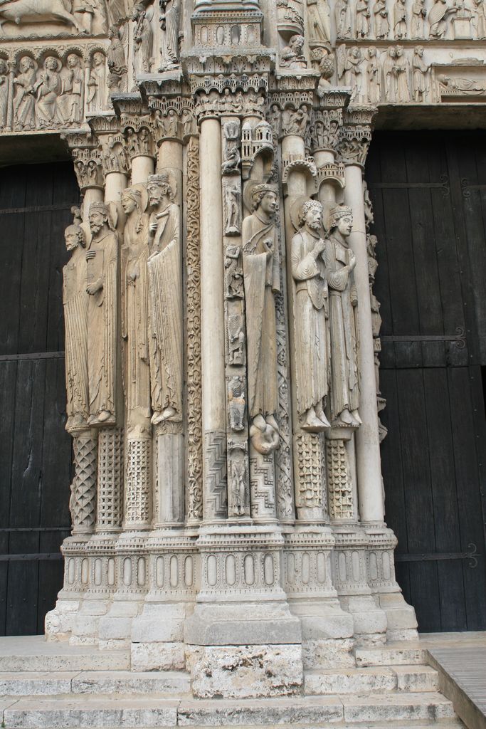 Elongated standing figures line decorated shafts between two dark doorways, below carved canopies.