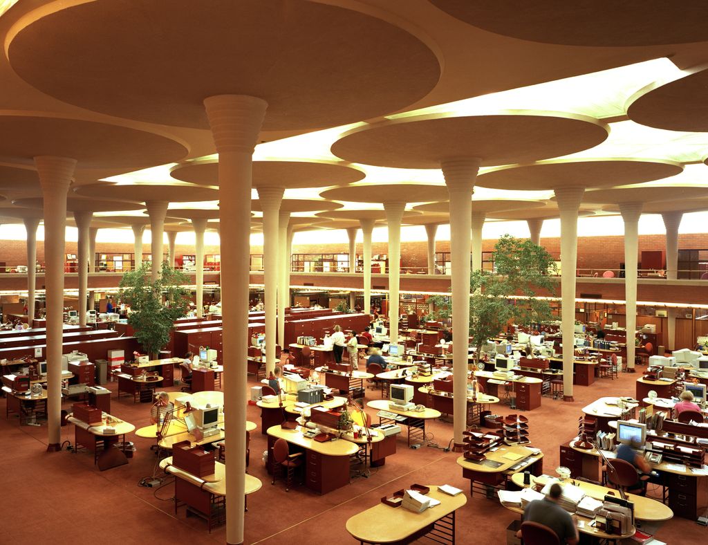 Slender columns broaden into circular roof-supporting pads above desks, plants and office workers.