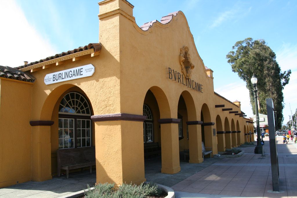 A long ochre station frontage repeats round arches and thick piers along a shaded pedestrian route beside modern paving, benches and signs.
