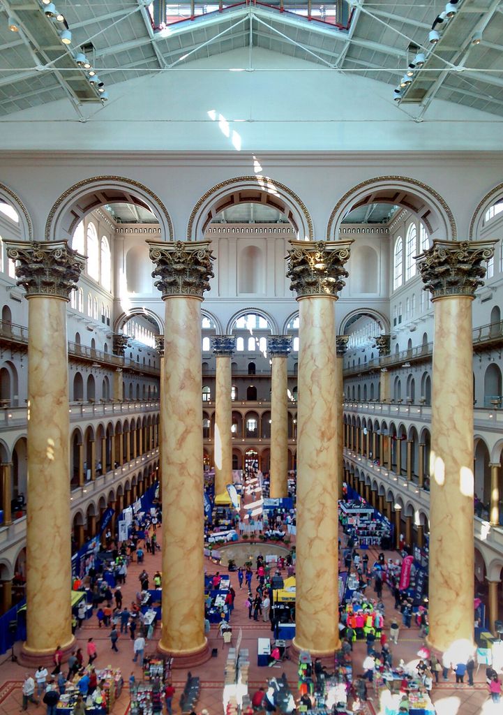An elevated interior view shows colossal painted columns, stacked arcades, high wall openings and roof glazing around a crowded central court.