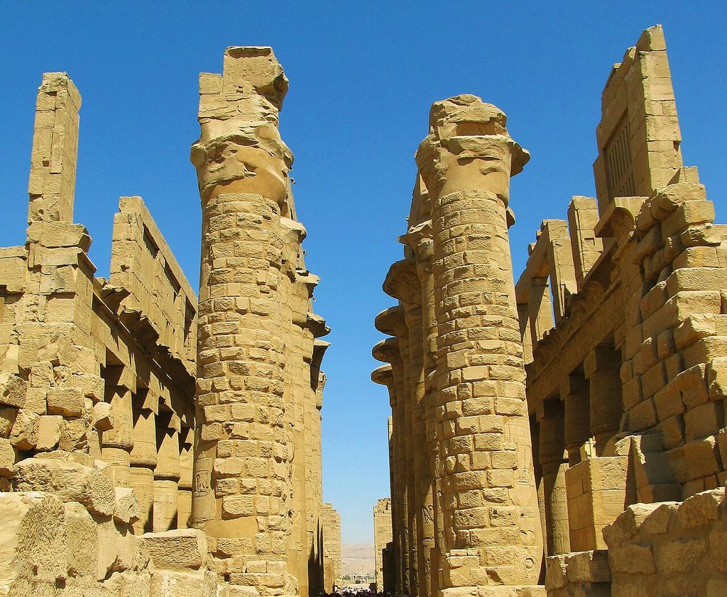 Massive carved stone columns and surviving high stone grille openings rise into an open blue sky where much of the ancient roof is missing.