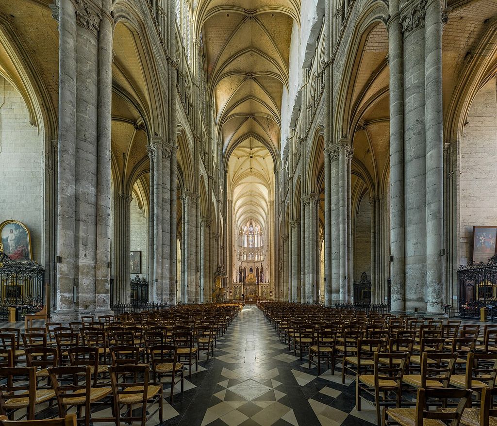 A long Gothic nave rises through pointed arcades and an intermediate wall zone to high windows and ribbed vaults above rows of modern chairs.