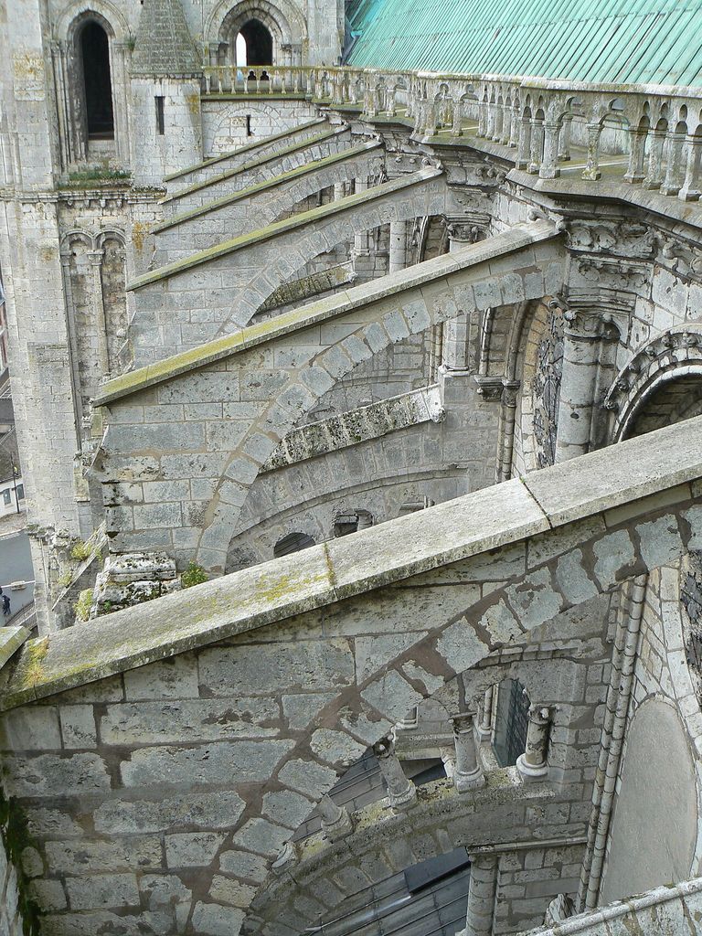 A roof-level view shows successive stone flying buttresses crossing above lower roofs toward the cathedral's upper wall, with joints, weathering and repairs visible.