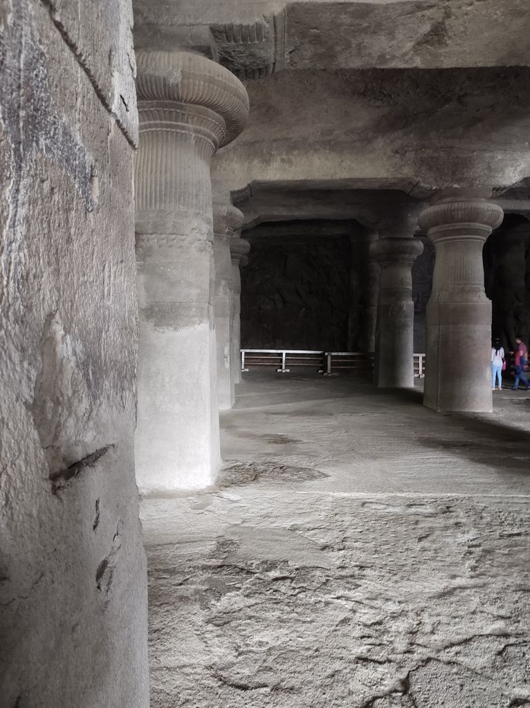 Rows of rock-cut stone pillars with fluted tops and rounded capitals recede through a dim cave hall beneath broad carved beam forms.