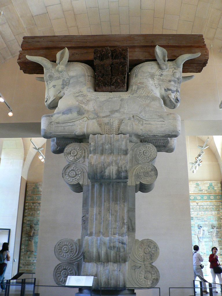Two carved bull foreparts face opposite directions above stacked stone scrolls and a fluted support, with dark timber beams displayed across their backs.