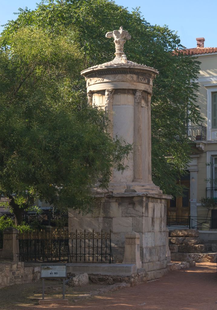 A round marble superstructure with slender Corinthian columns rises on a tall square podium among trees and city buildings.
