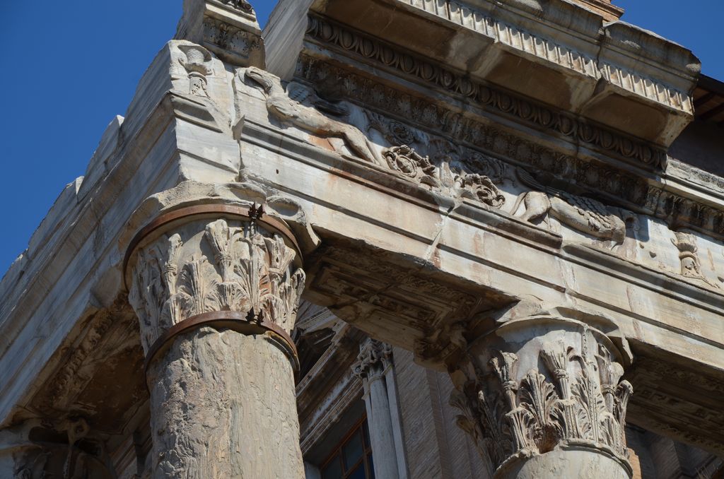 Two leafy capitals top weathered green-grey shafts beneath a carved Roman entablature, with later church fabric behind.