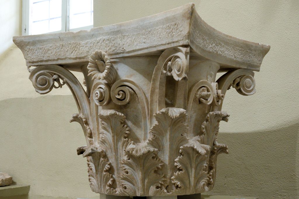 A large marble capital has two tiers of deeply carved leaves, rising stalks, spiral corner forms and a curved square abacus.