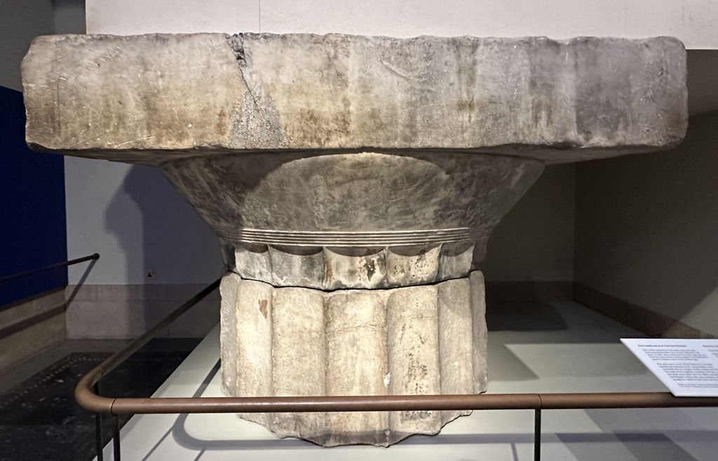 A detached marble Doric capital and fluted top drum stand behind a low rail in a museum gallery.