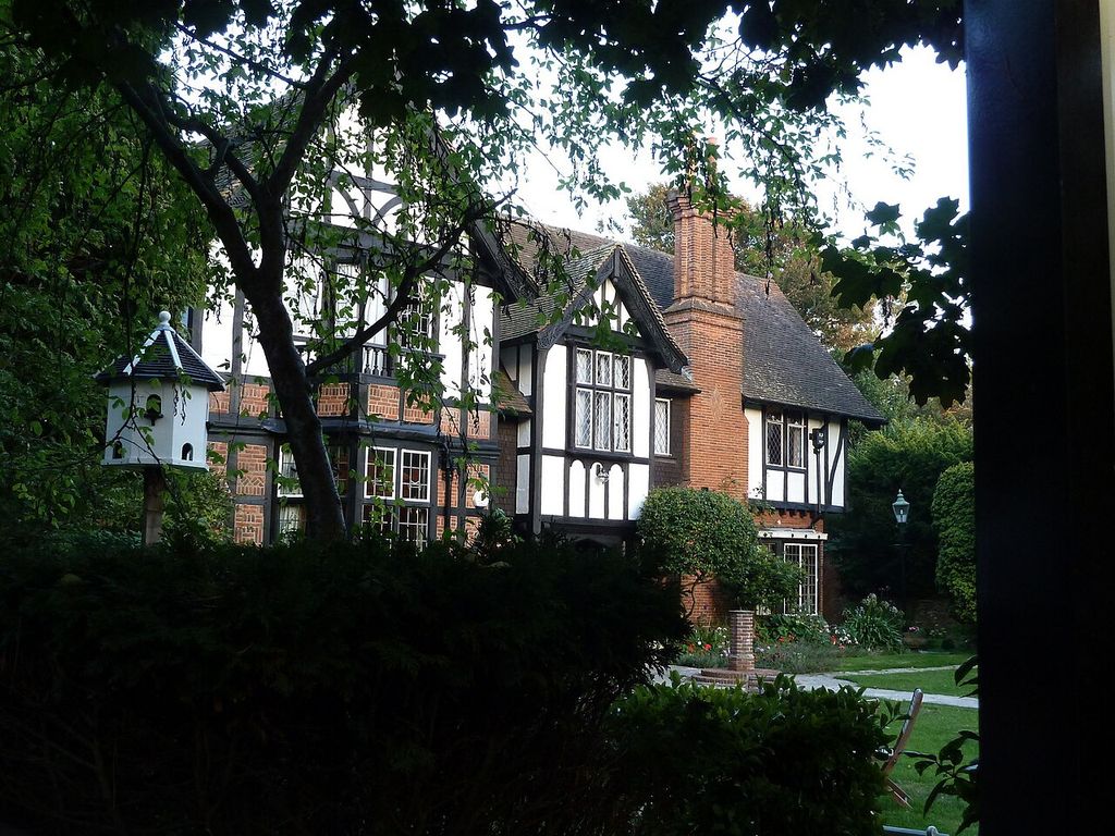 A large brick-and-white house with applied dark timbering, projecting bays, steep gables and tall chimneys appears through dense garden foliage.