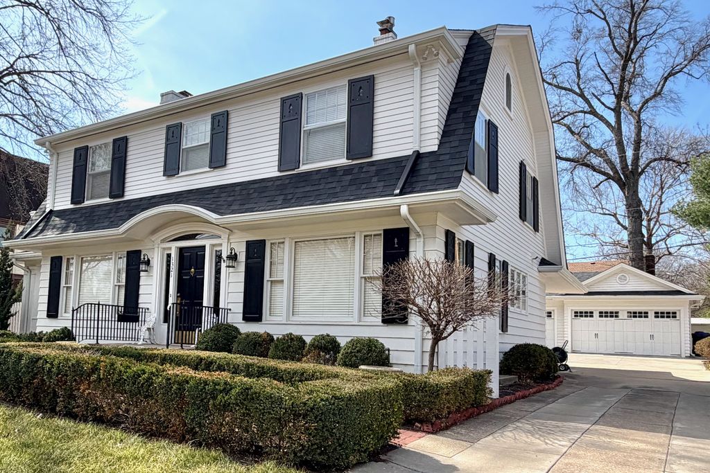 A white suburban house has a broad two-slope gambrel roof, upper dormer windows, black shutters and a separate gabled garage.