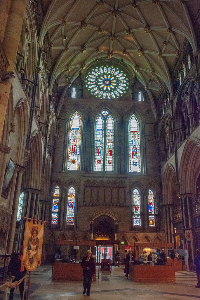 A luminous rose and three tall lancets rise in a vaulted transept wall above visitors, furnishings and a staffed interior area.