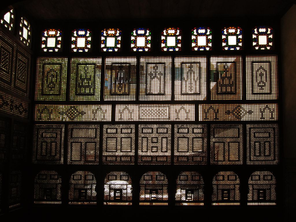 Daylight passes through dense geometric wooden lattice panels beneath a row of small coloured-glass windows.
