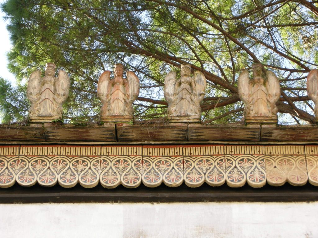 Painted palmette tiles and repeated winged figures line a timber roof edge beneath green pine branches.
