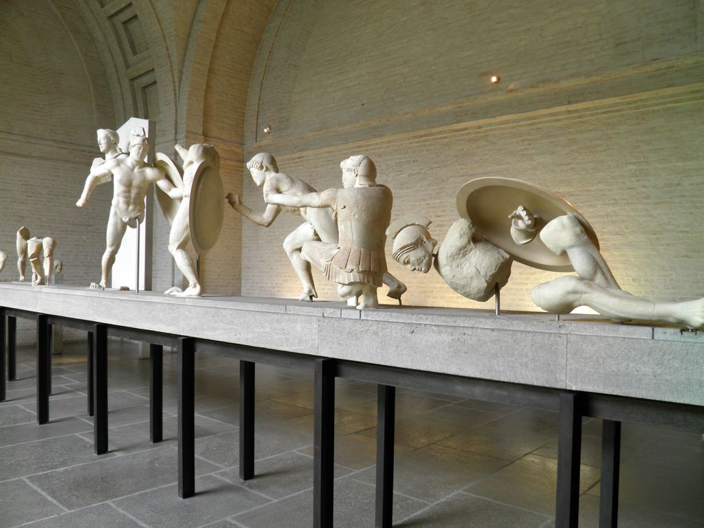 A museum row of marble warriors shifts from standing figures toward a crouching and reclining warrior at the low right end.
