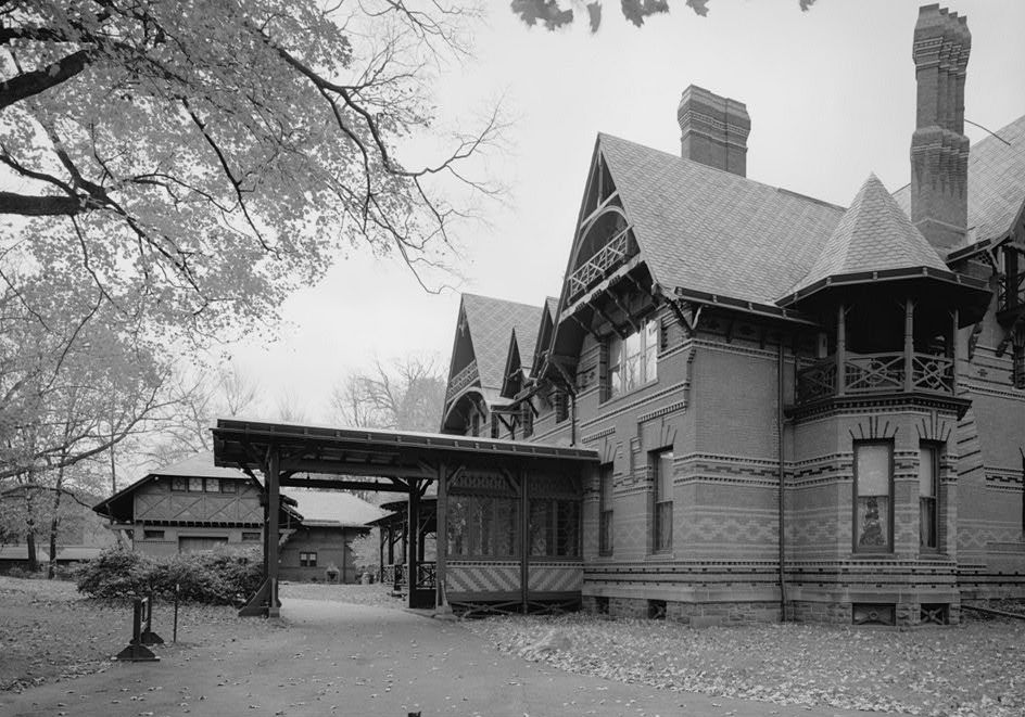 A long open timber canopy crosses a driveway between a patterned brick house and a lower carriage house in a black-and-white survey photograph.