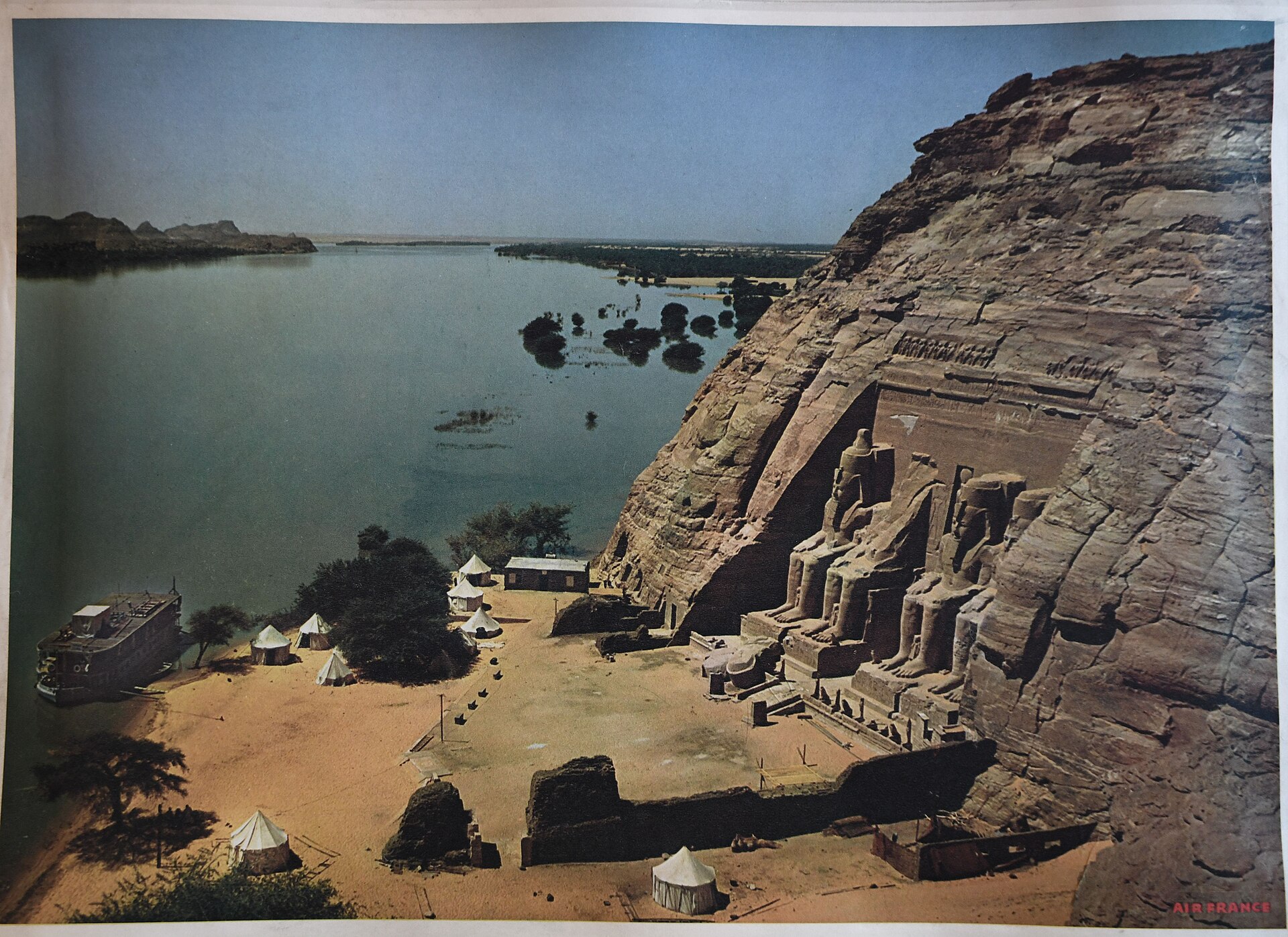 Historic elevated colour view of the Great Temple beside the close Nile, with a riverboat, trees, tents and camp at its foot.