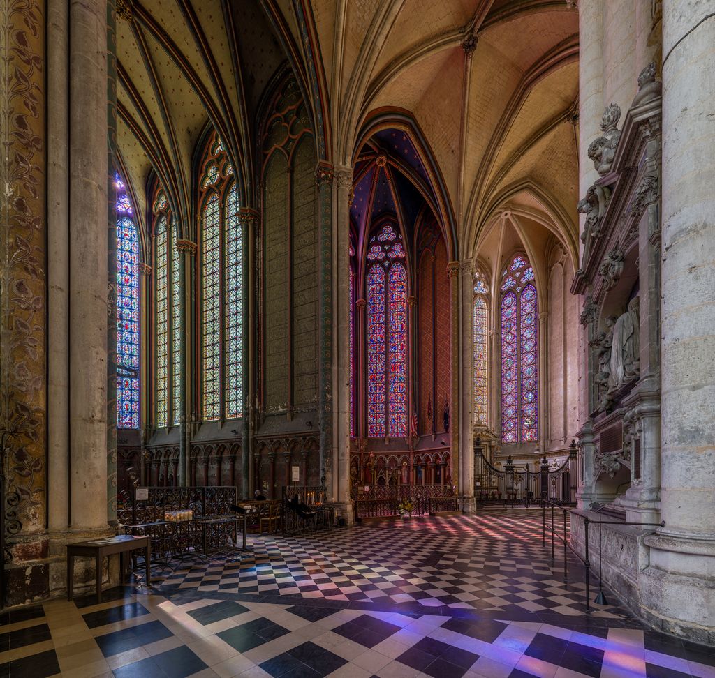 A vaulted cathedral ambulatory curves past tall coloured chapel windows, slender columns, patterned paving, railings and a large wall monument.