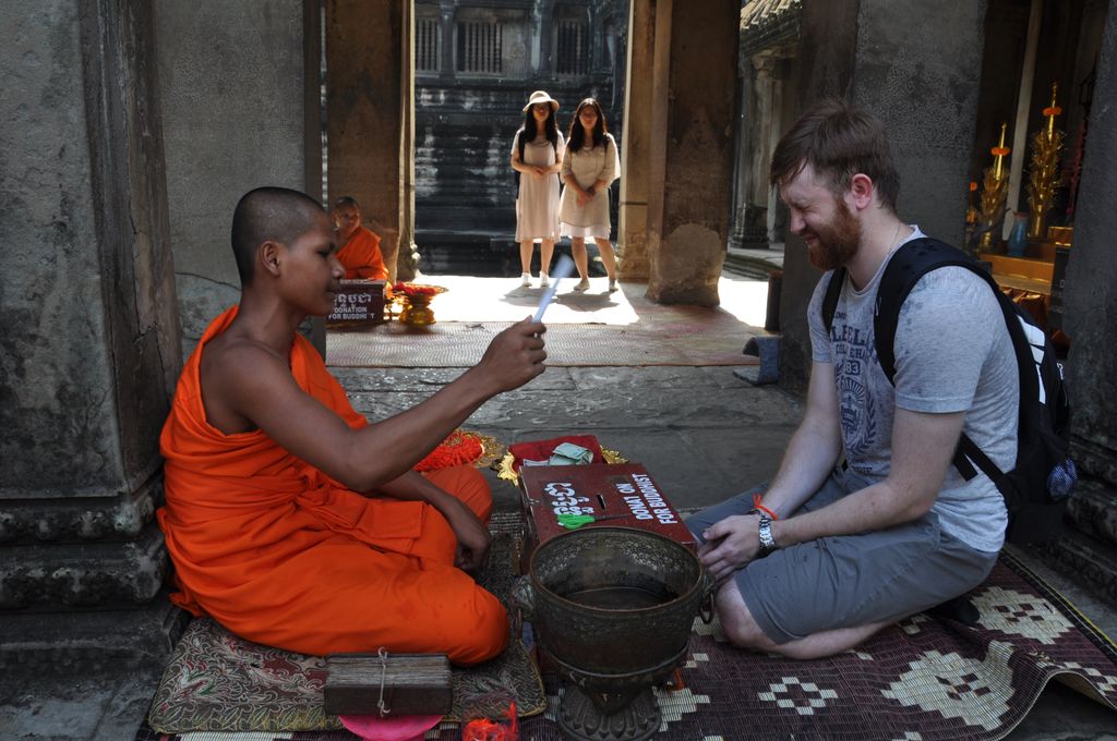 A seated monk in bright orange robes gives a thread blessing to a kneeling visitor on woven mats inside a dark stone temple bay.