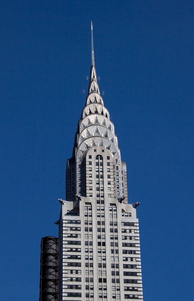 A tall Art Deco skyscraper rises against dark blue sky through brick setbacks, projecting eagles, seven bright metal crown arches and a slender needle.