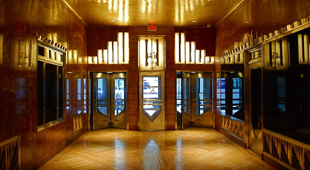 A wide symmetrical Art Deco hall glows amber around metal-and-glass doors, polished red-brown walls, stepped light panels and a geometric wood floor.