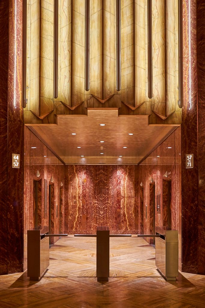 A richly finished elevator hall of red marble, pale figured stone, bronze-toned strips and geometric wood is seen through three modern turnstiles.