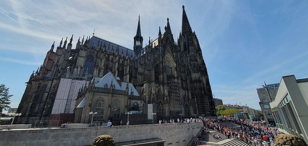 Cologne Cathedral rises above the station approach, with the twin west towers, transept, nave roof and patches of conservation scaffolding visible.