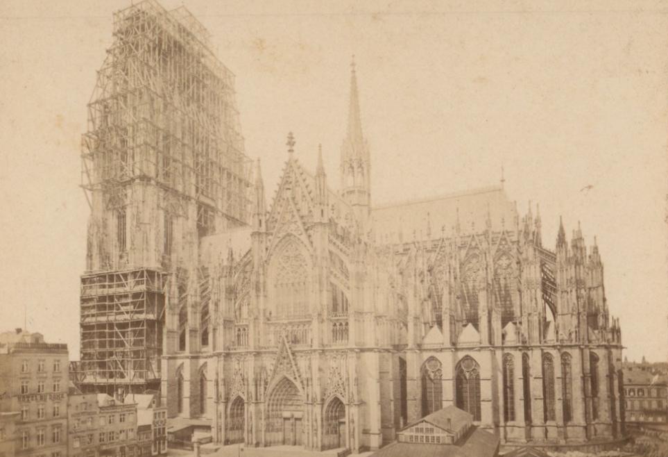 A sepia albumen photograph shows Cologne Cathedral from the south while enormous timber scaffolds surround the rising west towers.