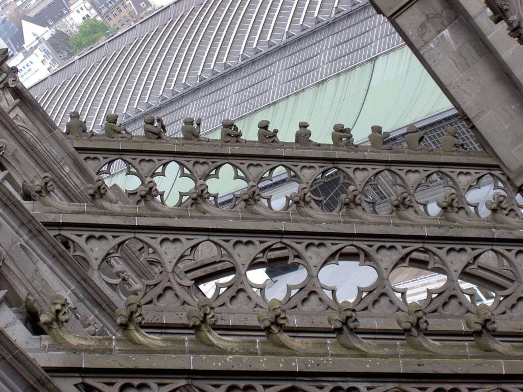 Traceried flying-buttress bridges cross between pinnacled outer piers and Cologne Cathedral’s high wall, with carved craftsman figures along the stonework.