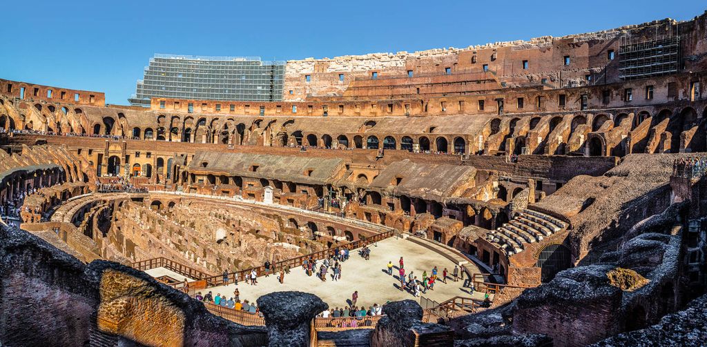 A wide interior panorama looks over the Colosseum's exposed underground walls and corridors toward broken seating tiers, arched passages, visitors and modern conservation structures.