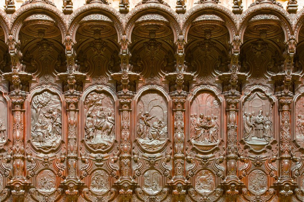Warm carved wooden choir stalls repeat small arches, columns and relief scenes in a richly ornamented horizontal wall.
