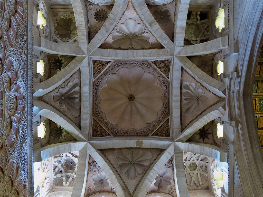 An ornate ceiling is divided by eight crossing pale stone ribs around a central octagon, with flower-like panels, stars and small windows.
