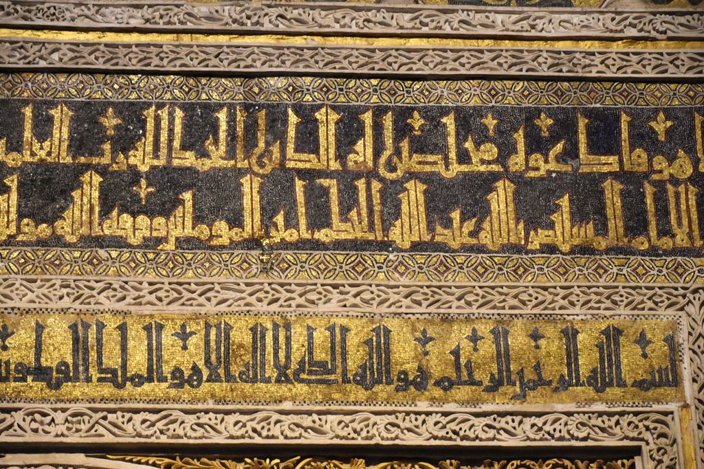 Two horizontal Arabic inscription bands in gold and dark mosaic are separated by intricate pale carved vegetal borders.
