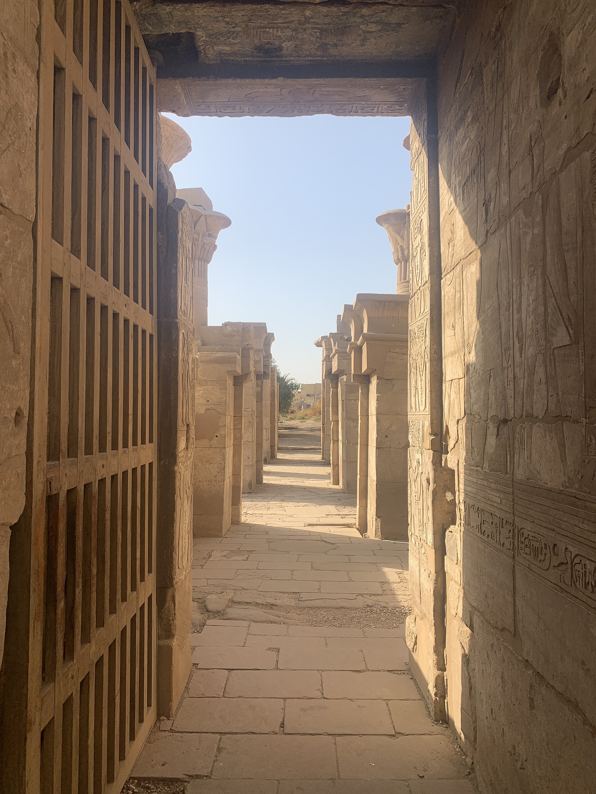 View through a roofed decorated stone doorway into the aligned courts of Ptah’s temple, with a present wooden barrier at left.
