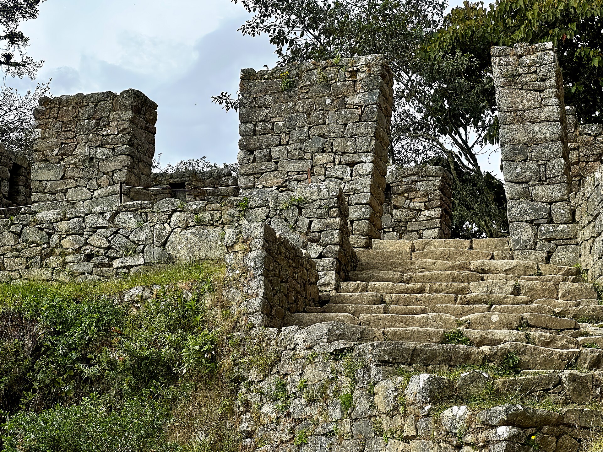 Intipunku's roofless Inca stone gateway and stairs amid grass and trees on the road approach.