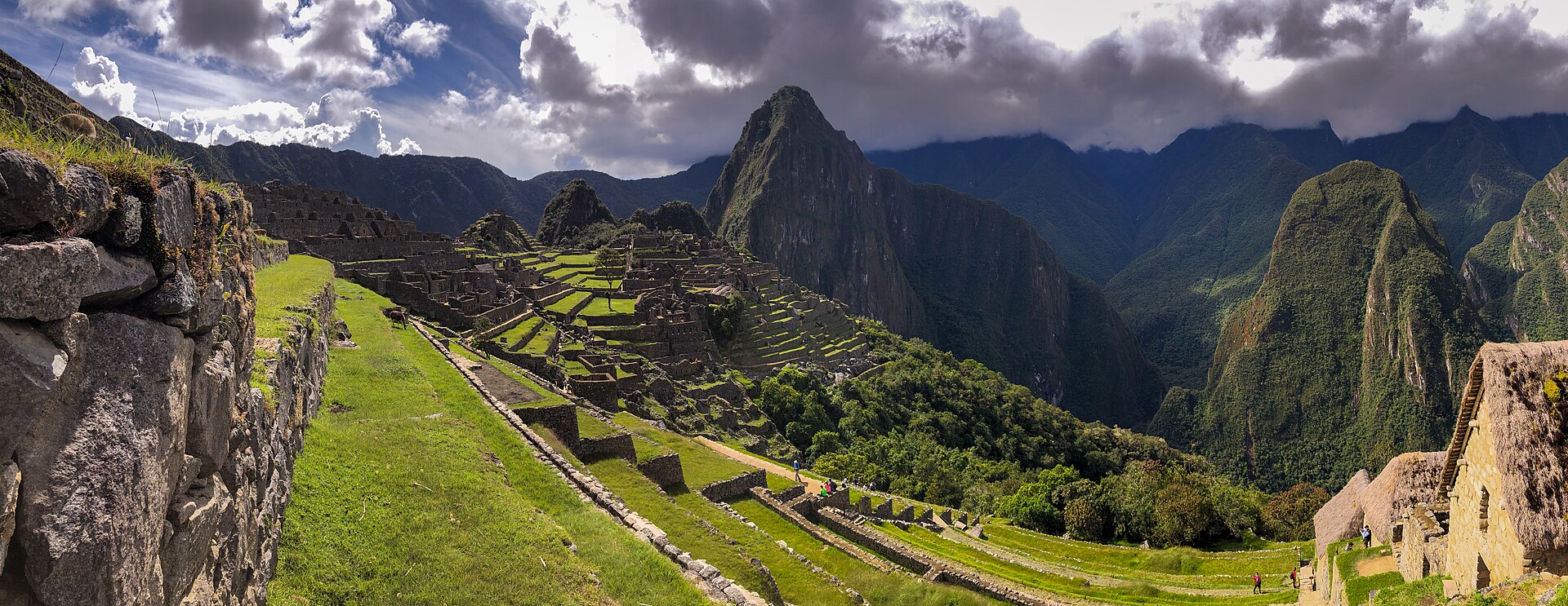 Wide ridge landscape with roofless Inca stone walls and green terraces below clouded mountain peaks.