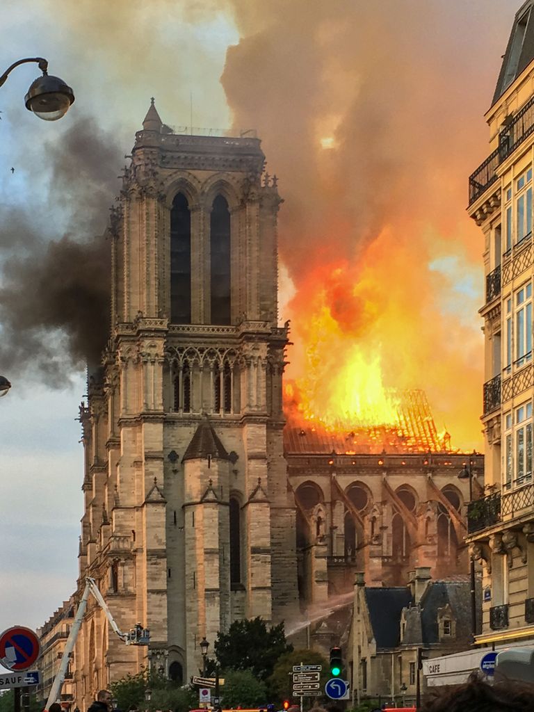 A cathedral tower stands beside an intensely burning roof, exposed timber framing, orange flame and dark smoke, while water streams rise from emergency vehicles below.