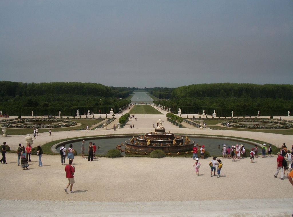 A fountain and formal flower parterres lead along a broad tree-lined axis toward a long rectangular canal on the horizon.
