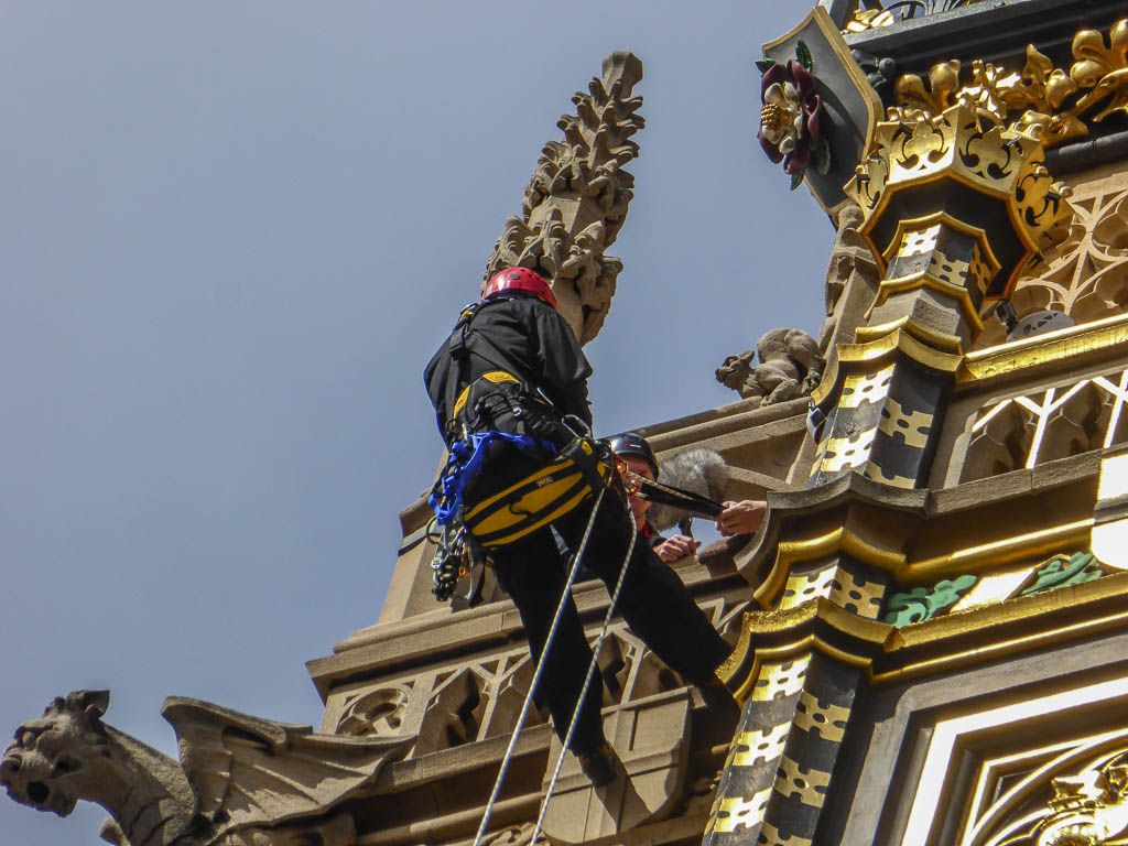 Two rope-access workers in helmets clean ornate gilded, painted and carved details high on a Gothic clock tower against blue sky.