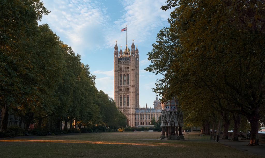 A tall square Gothic tower with pinnacles and a flag rises at the end of a long tree-lined lawn beside an ornate memorial fountain.
