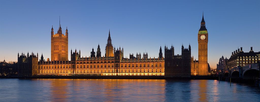 A long illuminated Gothic palace stretches beside a river at dusk, with a broad square tower at left, slender central spire and clock tower at right.