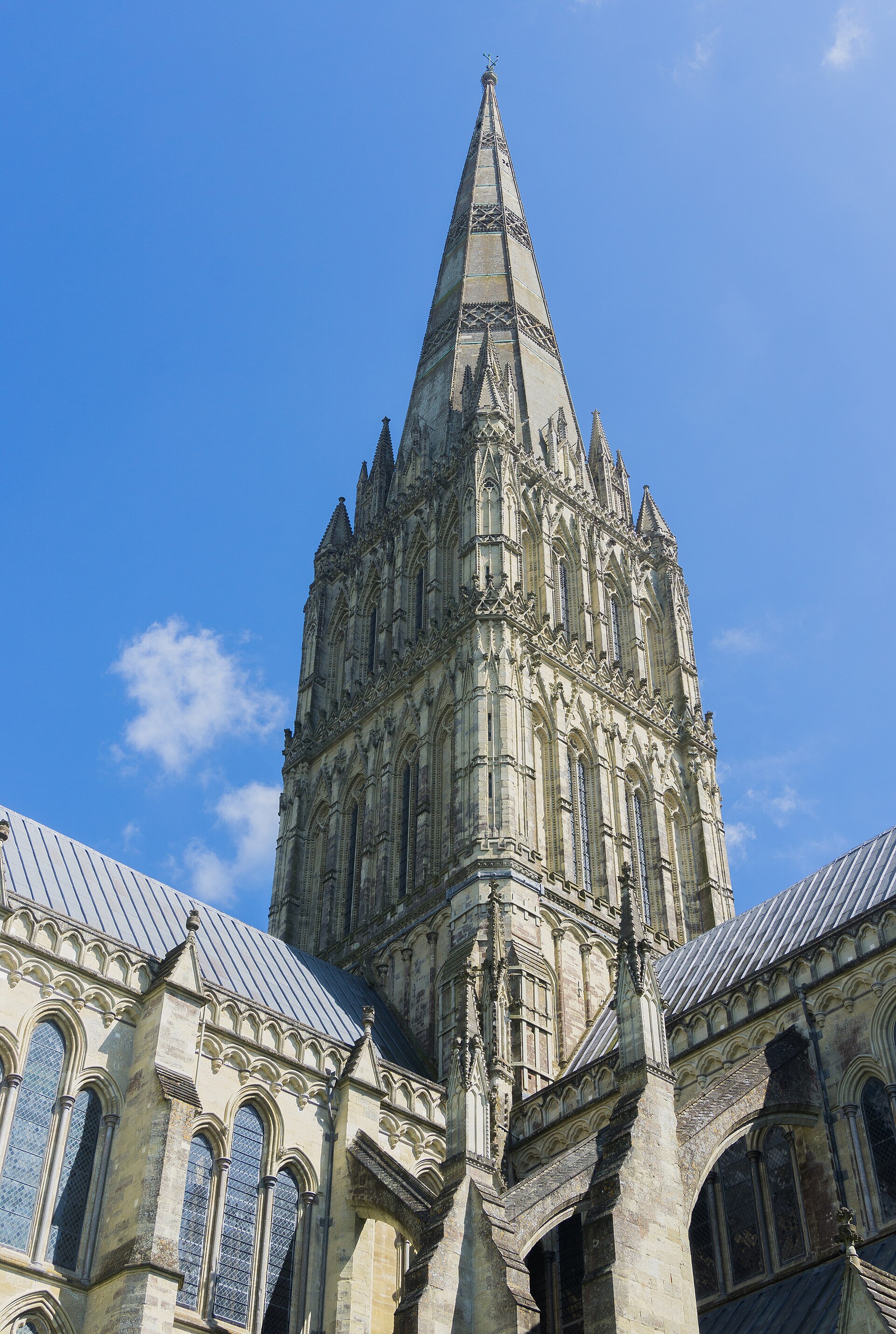 Salisbury Cathedral's pale stone tower and tall tapering spire rising above transept roofs and flying buttresses against a blue sky.