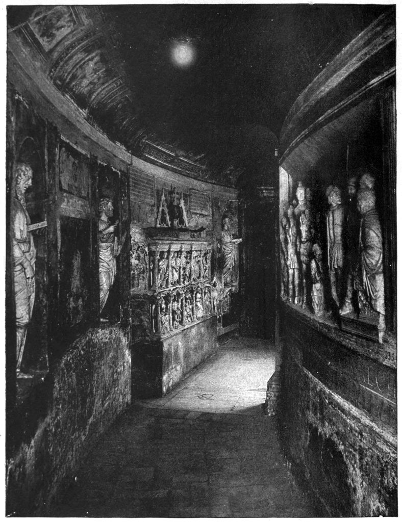 A dark historical photograph shows a narrow curving vaulted passage lined with tombs, reliefs and sculpted fragments beneath St Peter's Basilica.