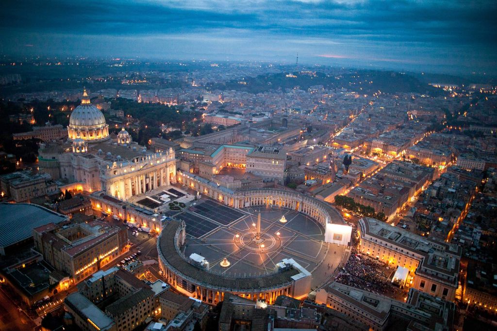 A twilight aerial view shows St Peter's Basilica and dome behind a trapezoidal forecourt and a vast oval colonnade around an obelisk and fountains.