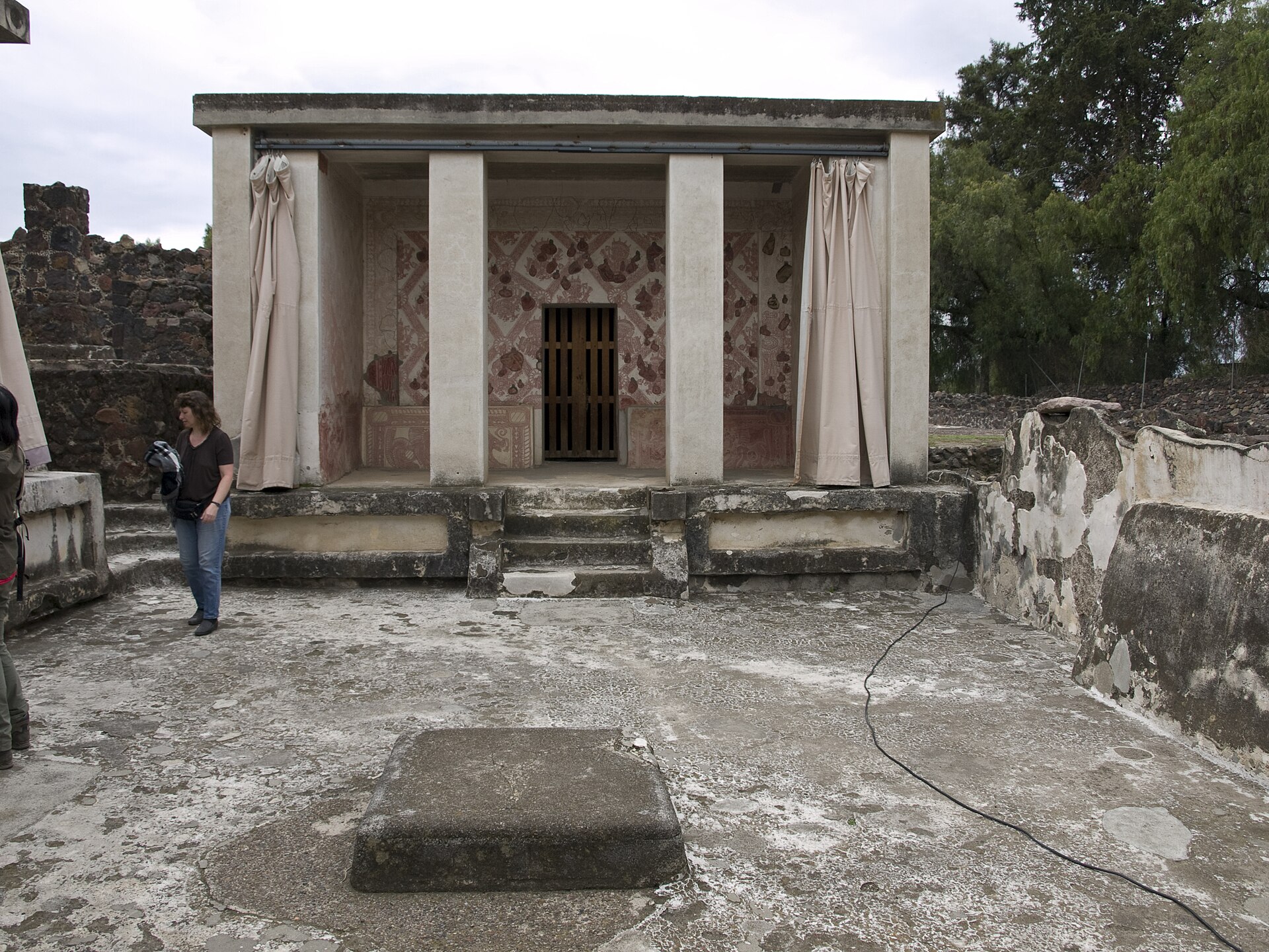 Small painted Atetelco portico with preserved red wall images, modern roof, pale curtains and visitor in foreground.