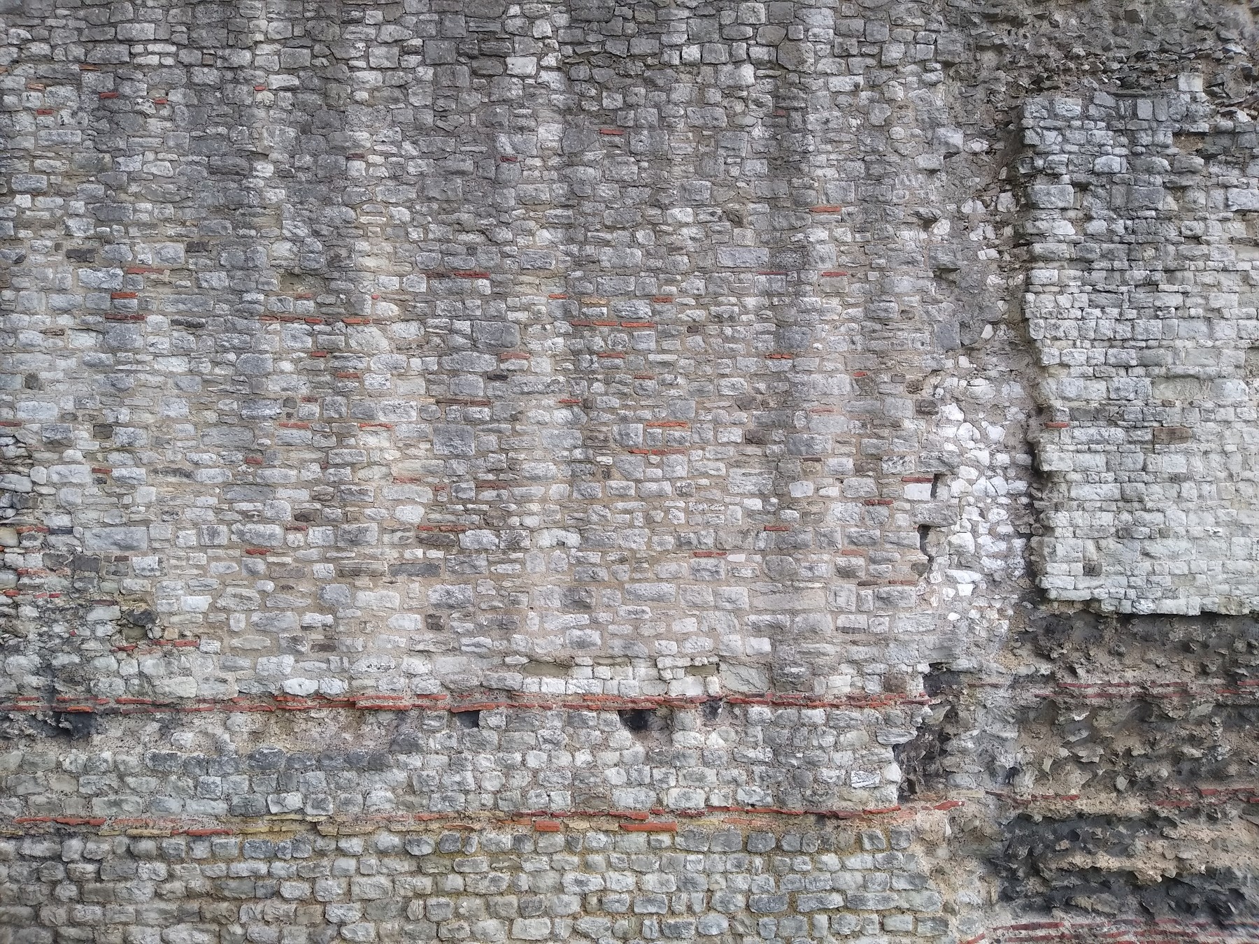 Rough stone and mortar wall with several horizontal bands of thin red ceramic tiles.