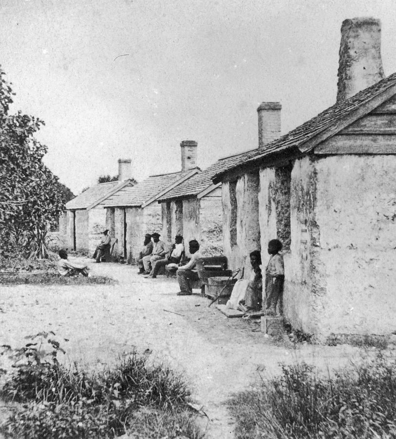 Historic monochrome view of a row of roofed cabins, with people and domestic objects beside the doorways.