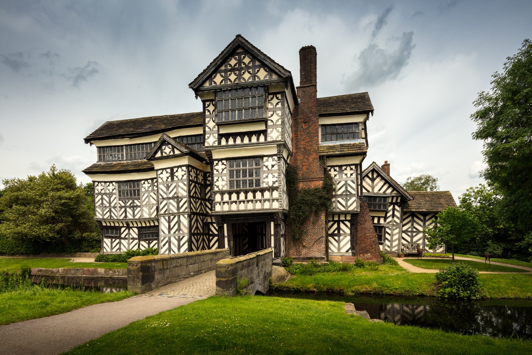 Little Moreton Hall across its stone entrance bridge, with dark patterned framing, pale panels, projecting upper walls and a tall brick chimney.