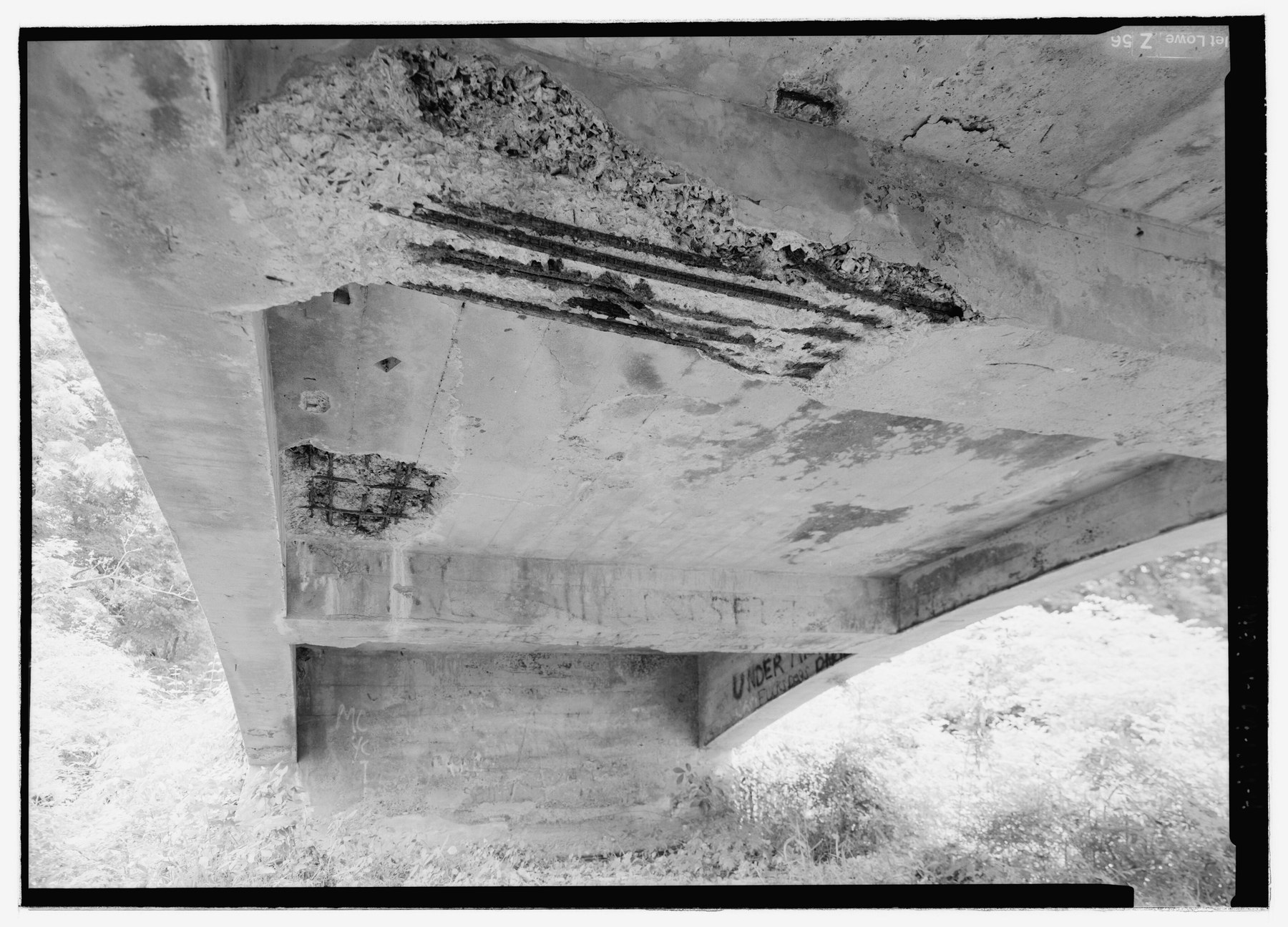 Black-and-white underside view of a ribbed concrete bridge with spalling and exposed reinforcing bars.