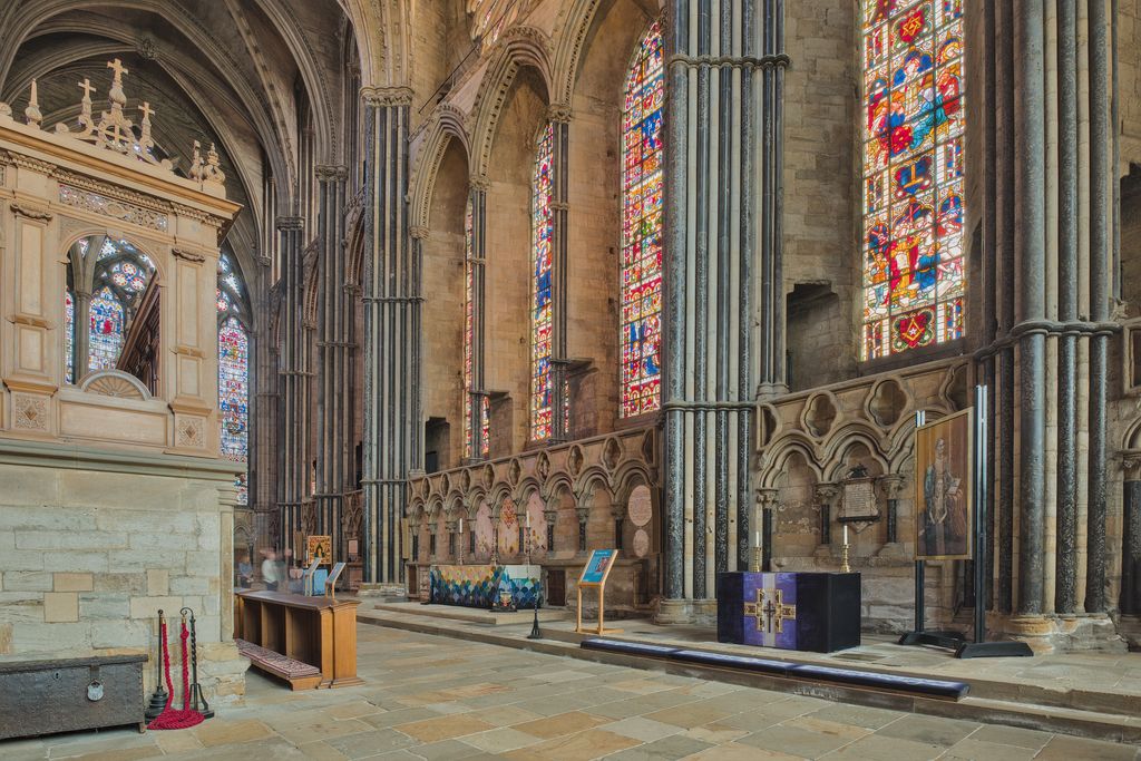 Altars stand beside a straight stone wall with tall stained-glass windows, clustered shafts and a lower floor beside a shrine enclosure.